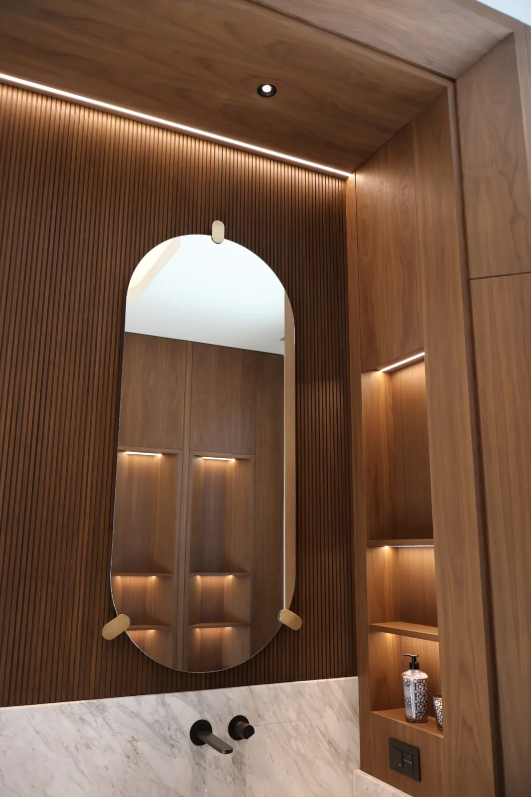 Arca Konakları — Master bathroom — cove light and lit niches set into fluted walnut frame a capsule mirror.