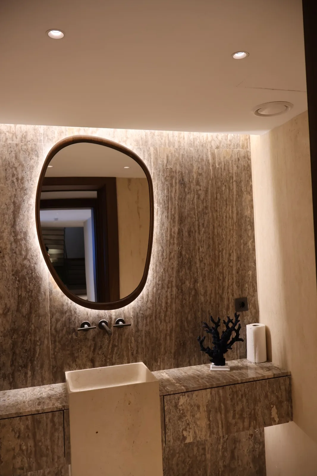 Arca Konakları — A guest powder room: recessed downlights and a back-lit mirror on book-matched travertine.